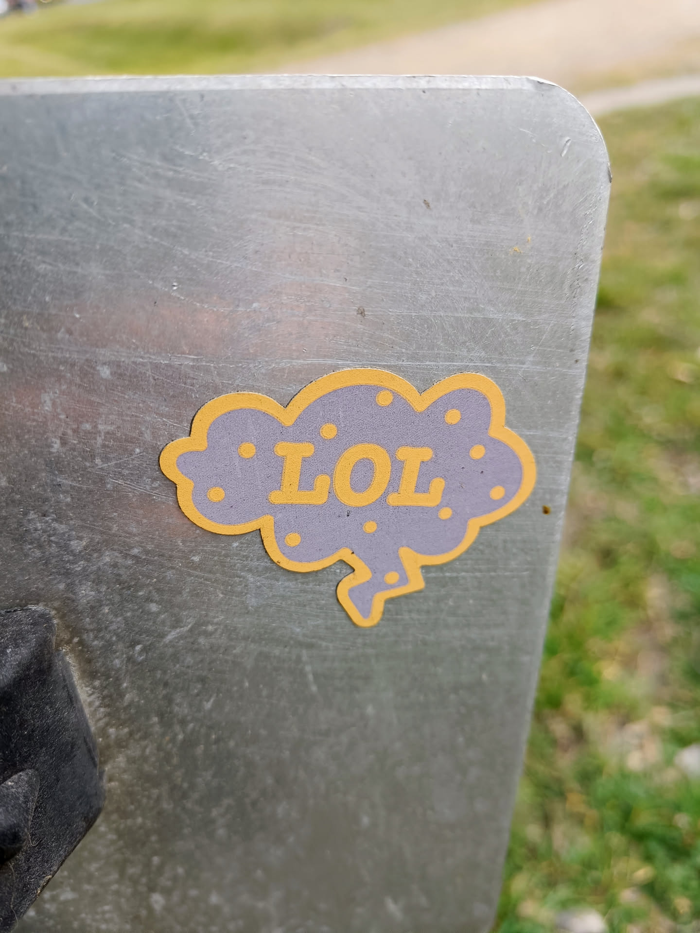 A small sticker of a purple cloud with the letters LOL in yellow. The sticker is on a scratched metal surface.