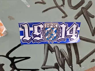 A blue and white sticker commemorating the year 1914 and featuring the logo of IFK, possibly a sports club or organization.  The logo includes a lion emblem.