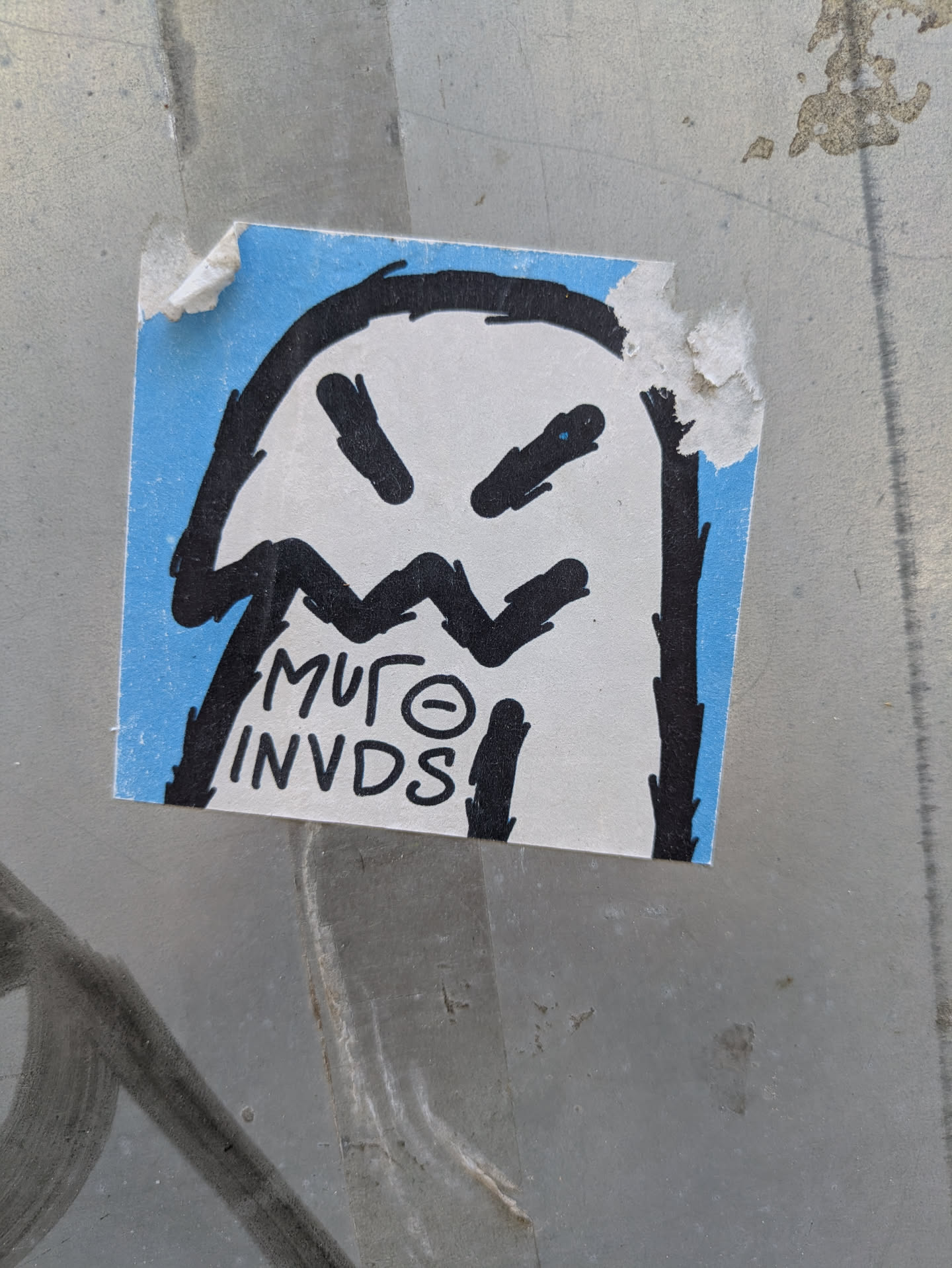 A blue and white square sticker features a ghost-like character with angry black eyes and a jagged mouth. The style is reminiscent of graffiti art with thick black outlines. Text below the character reads 'MUFO INVDS'. The sticker is adhered to a gray surface.