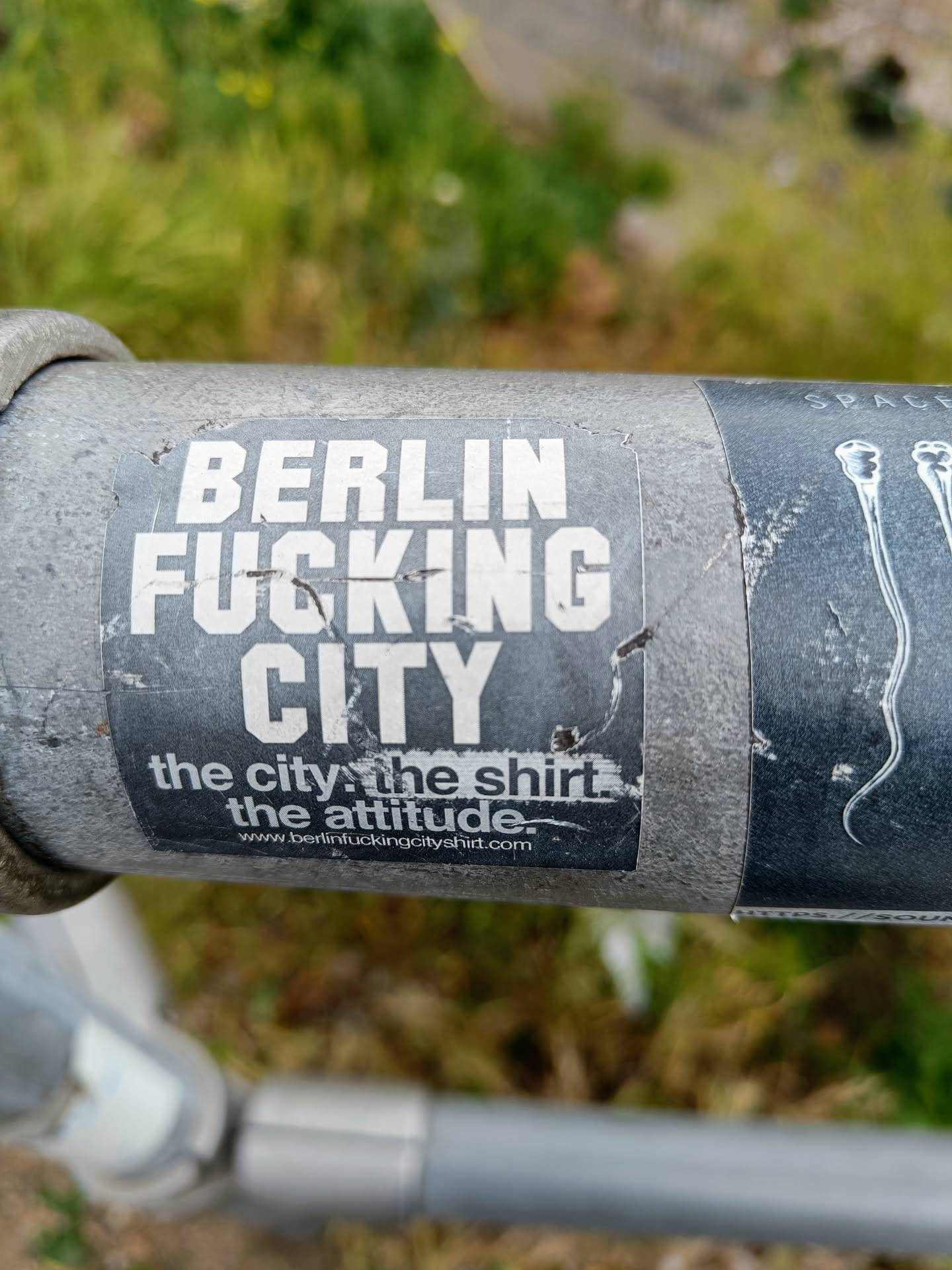 A worn sticker on a gray metal pole.  The sticker features the text "BERLIN FUCKING CITY" in large, bold letters, with the tagline "the city, the shirt, the attitude." and a website address below. A portion of another sticker is visible to the right.
