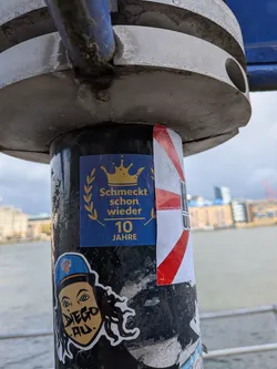 A rectangular sticker featuring a dark blue background. At the top, a golden crown is prominently displayed. Below the crown, a golden laurel wreath frames the German text 'Schmeckt schon wieder'. Further below, the number '10' is written in a larger golden font, followed by 'JAHRE' (years) in smaller golden text. The sticker has a celebratory and regal theme, with a clean design in blue and gold.