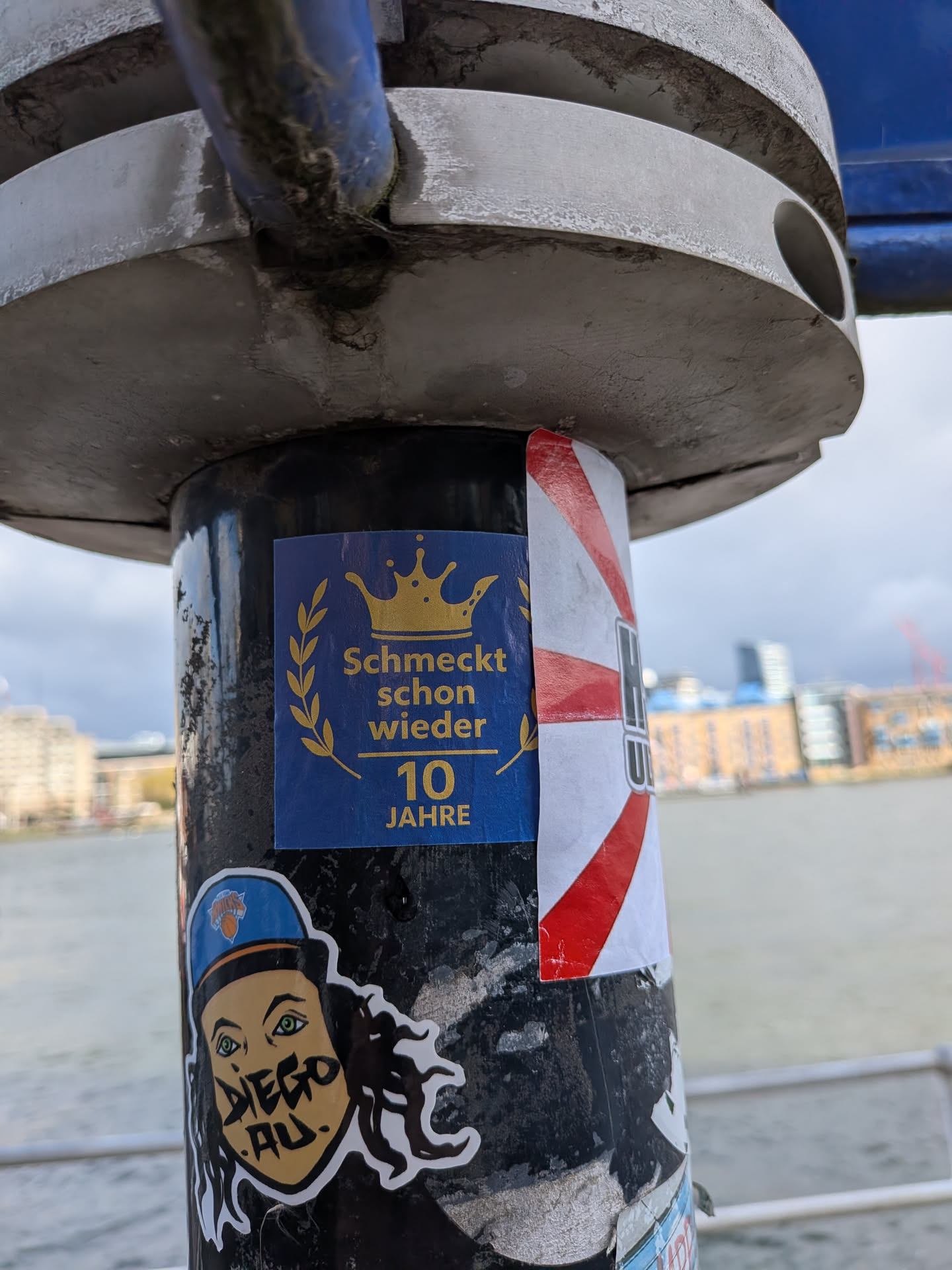 A rectangular sticker featuring a dark blue background. At the top, a golden crown is prominently displayed. Below the crown, a golden laurel wreath frames the German text 'Schmeckt schon wieder'. Further below, the number '10' is written in a larger golden font, followed by 'JAHRE' (years) in smaller golden text. The sticker has a celebratory and regal theme, with a clean design in blue and gold.