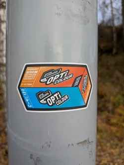 The sticker is hexagonal with rounded corners and a white border. It is divided into two main sections: the top half is orange and the bottom half is blue. The top section features text in white and orange, including 'VIBRANT', 'WARM', 'DETAILED', 'VERSATILE', and 'CONTRASTY'. Below this, in a smaller orange box, is 'C-41/CN16'. The top right of the orange section has a logo with 'OPTIK OLDSCHOOL' above 'OPTI COLOUR' in bold black and white lettering, with a stylized representation of a film reel. The bottom blue section has 'C-41/120' in white text on the left. The bottom right of the blue section has a similar logo with 'OPTIK OLDSCHOOL' above 'OPTI COLOUR'. There are also numerical indicators on the right side of the sticker: '120', 'ISO', '200' in the orange section, and '120 ISO 200' in the blue section, suggesting film speed and format.