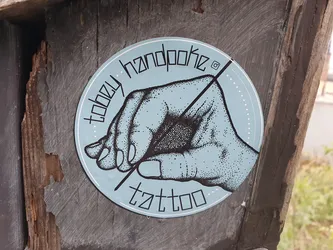 A light blue round sticker with a handpoke tattoo design. It features a hand holding a needle, with the text "Toozy Handpoke Tattoo" arranged around it.  A small Instagram icon is visible near the top.