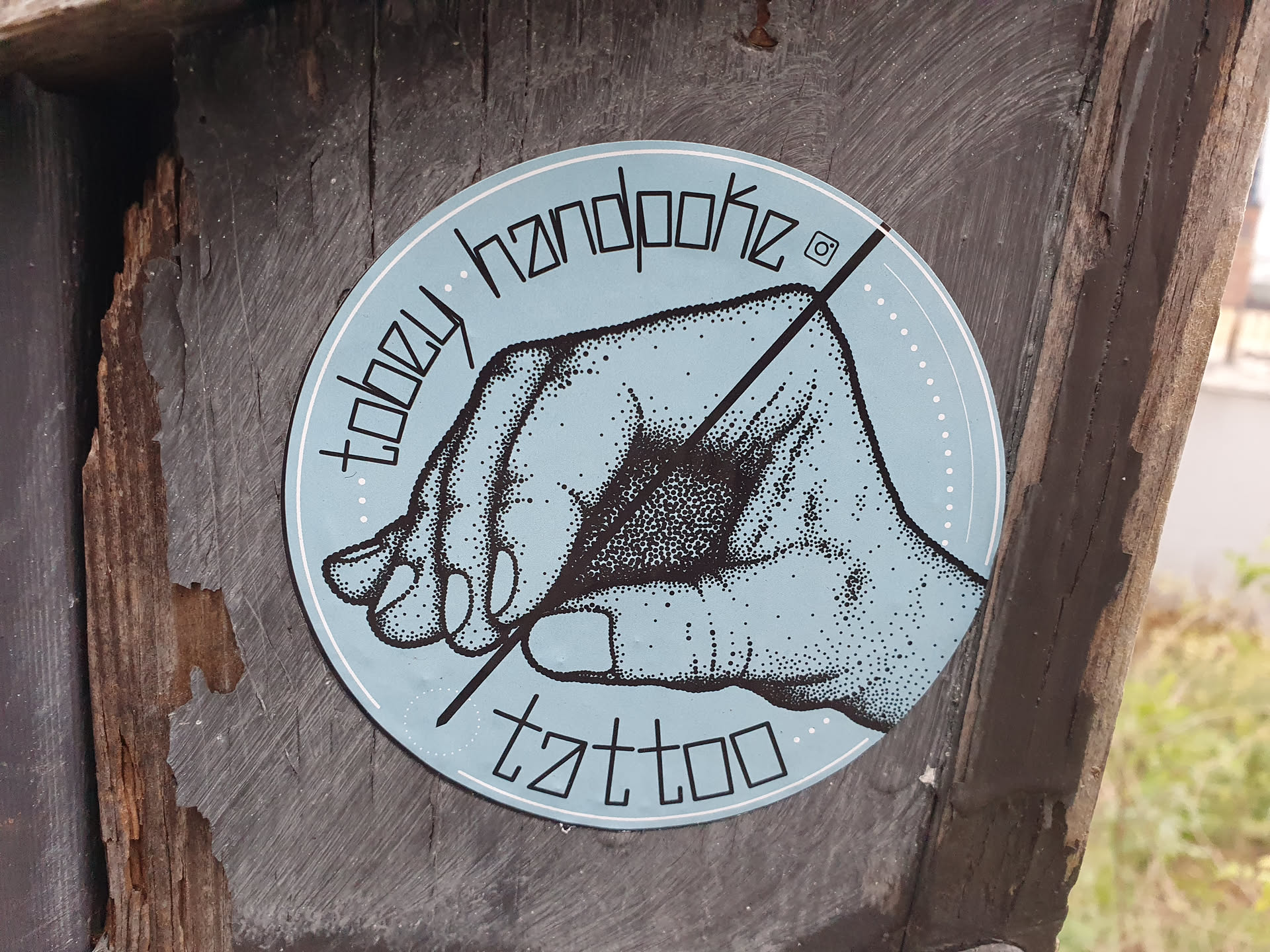 A light blue round sticker with a handpoke tattoo design. It features a hand holding a needle, with the text "Toozy Handpoke Tattoo" arranged around it.  A small Instagram icon is visible near the top.