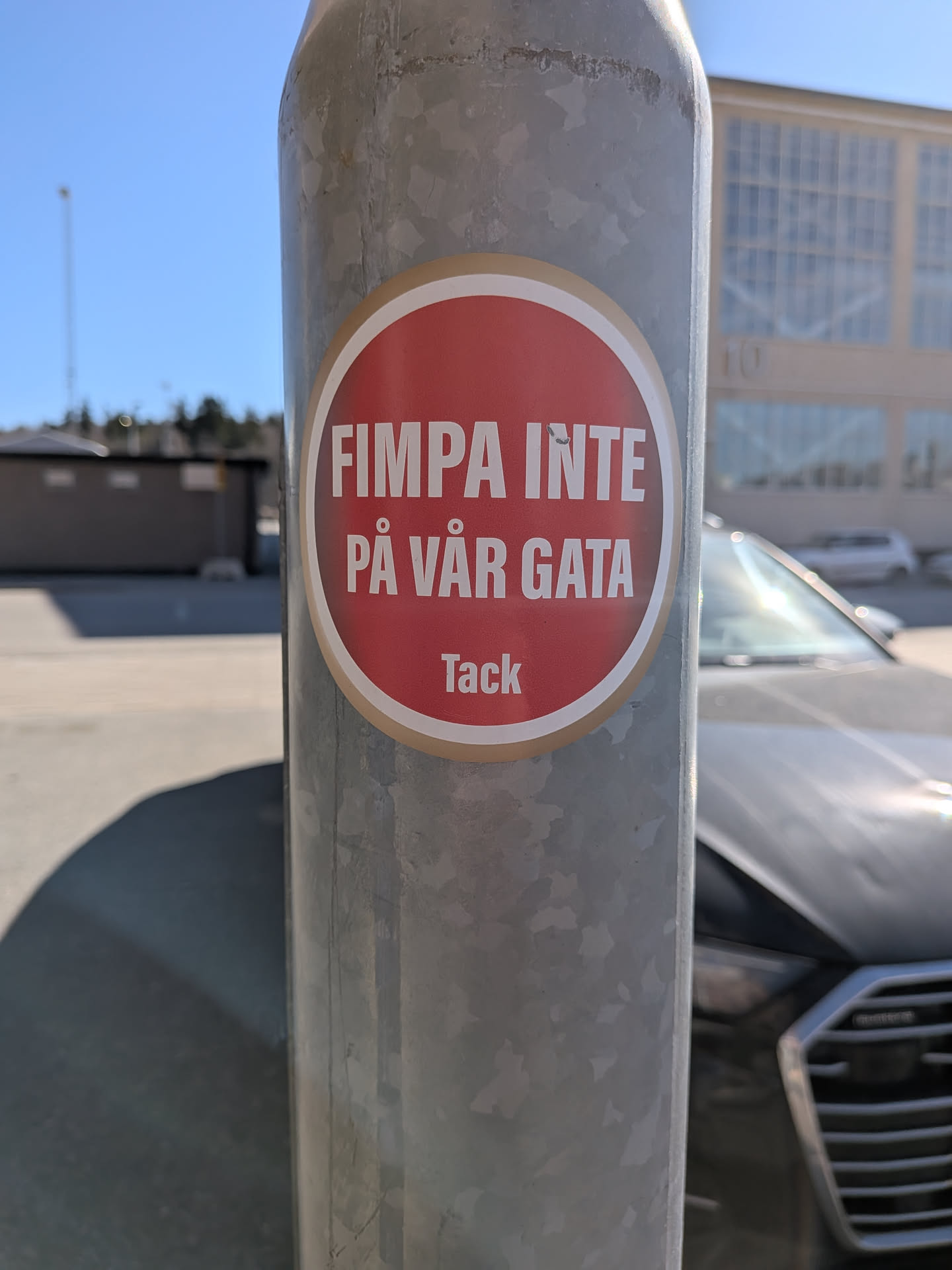 A circular sticker with a vibrant red background, featuring a thin white inner border and a slightly wider, light brown/beige outer border. The main text, 'FIMPA INTE PÅ VÅR GATA', is prominently displayed in bold, white, sans-serif capital letters across two lines. Below this, in smaller white sans-serif letters, is the word 'Tack'. The sticker is affixed to a textured, galvanized metal pole, with some minor wear visible around its edges.