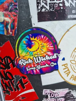 A tie-dye sticker with a sun and moon design. The text "Rick Wicked's Long Beach, Ca" is written on the sticker.