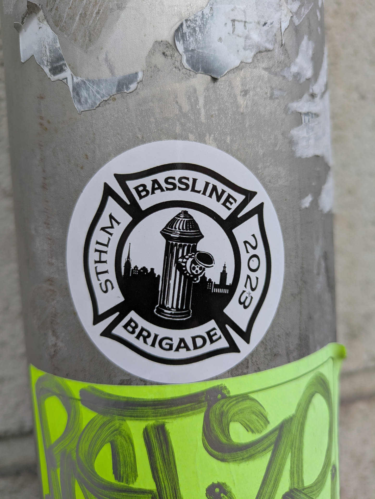 A round, white sticker affixed to a metal pole displays the text "BASSLINE BRIGADE STHLM 2023" arranged around a black emblem resembling a Maltese cross. The emblem features a fire hydrant in the center, set against a skyline. Below the sticker, there's some green graffiti art.