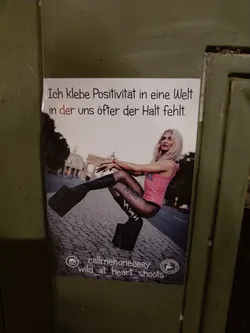 A sticker featuring a photo of a blonde woman in platform boots, sitting on a cobblestone street in front of the Brandenburg Gate. The text on the sticker translates to: "I stick positivity into a world where we often lack support."