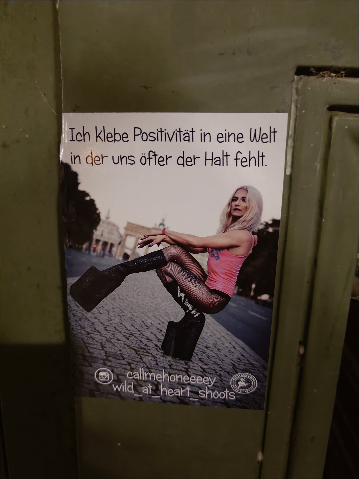 A sticker featuring a photo of a blonde woman in platform boots, sitting on a cobblestone street in front of the Brandenburg Gate. The text on the sticker translates to: "I stick positivity into a world where we often lack support."