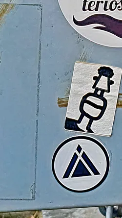 This image features a collection of stickers attached to a weathered, possibly metallic, surface. The most prominent sticker in the center is a white rectangle with a stylized black drawing of what appears to be a figure. Below it, there's a round sticker with a black triangle pointing upwards, surrounded by a black circle and filled with a gradient. The stickers seem to be part of a larger collection or a surface used for displaying various artworks.