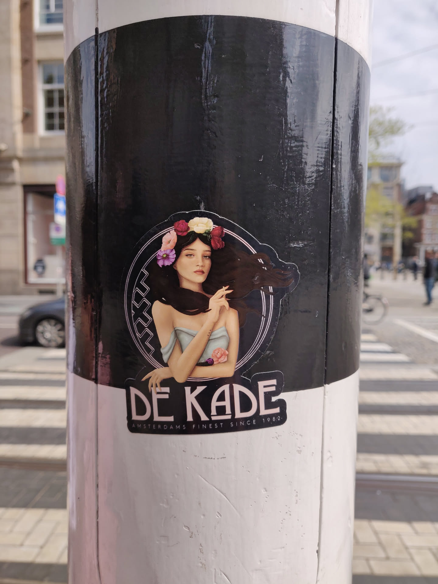 Sticker featuring a stylized illustration of a woman with flowers in her hair. The sticker is black and white with pink accents. It also contains the text 'DE KADE' and 'AMSTERDAMS FINEST SINCE 1986'.