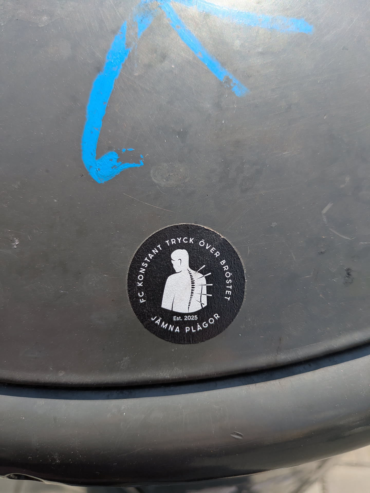 The sticker is black and circular, with white text and a white graphic. The central graphic is a minimalist silhouette of a human torso viewed from the back, highlighting the spine. Protruding from the spine are multiple short, sharp objects resembling needles or pins. Around the top curve of the circle, the text "FC KONSTANT TRYCK ÖVER BRÖSTET" is written in white capital letters. Around the bottom curve, the text "JÄMNA PLÅGOR" is visible, also in white capital letters. Below the central figure, a smaller text 'Est. 2025' is present. The design has a somber or unsettling tone, associating the imagery with the textual phrases.