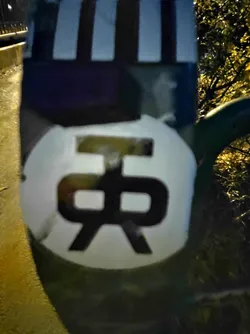 A circular sticker with a stylized black graphic on a white background. The graphic is bold and slightly abstract.  The sticker is affixed to a dark-colored surface with visible vertical stripes in the background. The photo was taken at night.