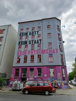 A building with a large mural on its facade. The mural displays the words "Kein Gott, Kein Staat, Kein Patriarchat" which translates to "No God, No State, No Patriarchy" in German.  The building shows other graffiti as well.