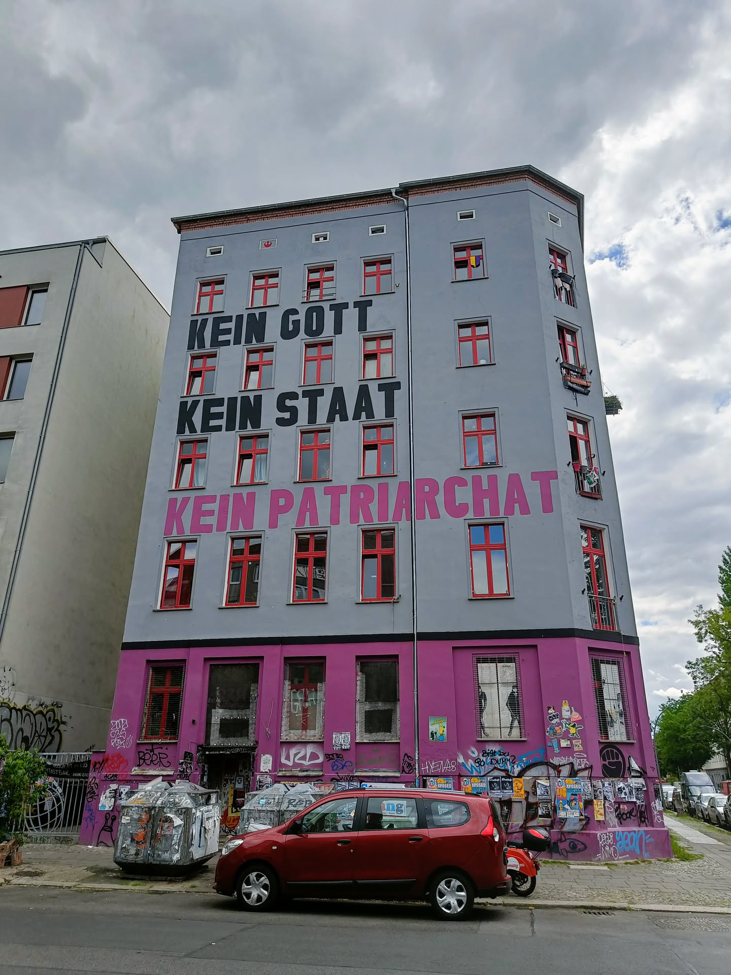 A building with a large mural on its facade. The mural displays the words "Kein Gott, Kein Staat, Kein Patriarchat" which translates to "No God, No State, No Patriarchy" in German.  The building shows other graffiti as well.