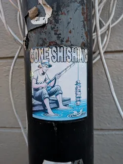 The sticker features a man wearing a hat, t-shirt, and shorts, sitting on a wooden pier with his feet in the water. He is holding a fishing rod, and the line is attached to a vertical stack of donuts. The water is depicted with ripples and splashes. The text 'GONE SHISHING' is prominently displayed at the top of the sticker in bold, white lettering with a black outline. The overall color scheme is vibrant, with blues for the water and sky, and muted tones for the man's clothing and the donuts.
