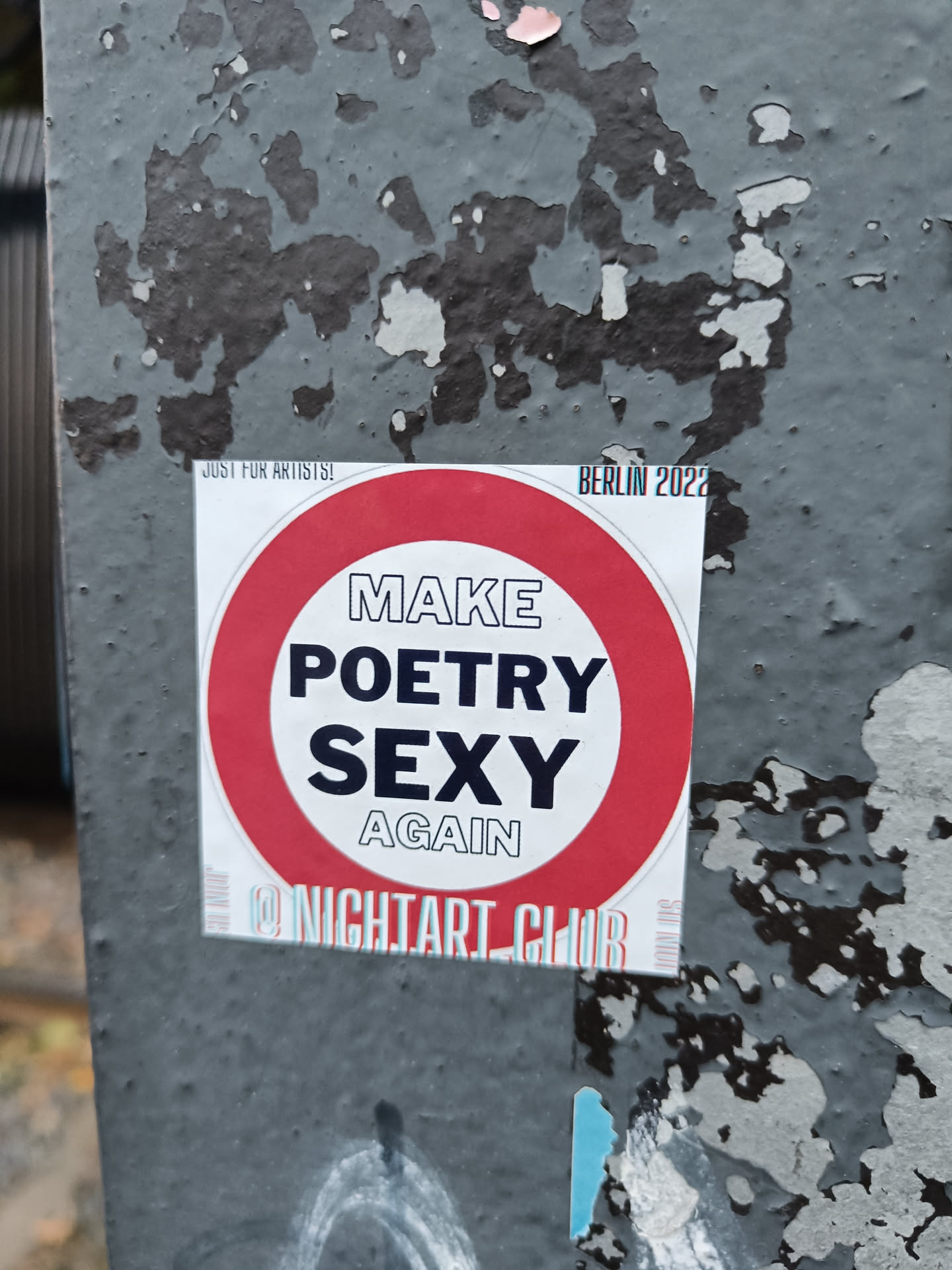 Round sticker with the text "Make Poetry Sexy Again" in the center.  It has a red circle border with white text. At the bottom it says @NightArtClub.  There is additional small text at the top that says "Just for Artists" and "Berlin 2022". The sticker is affixed to a weathered gray surface.