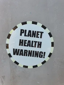A round sticker with a black and white design.  The text 'PLANET HEALTH WARNING!' is prominently displayed in bold, black capital letters on a white background. The sticker has a decorative border of alternating black and off-white segments.