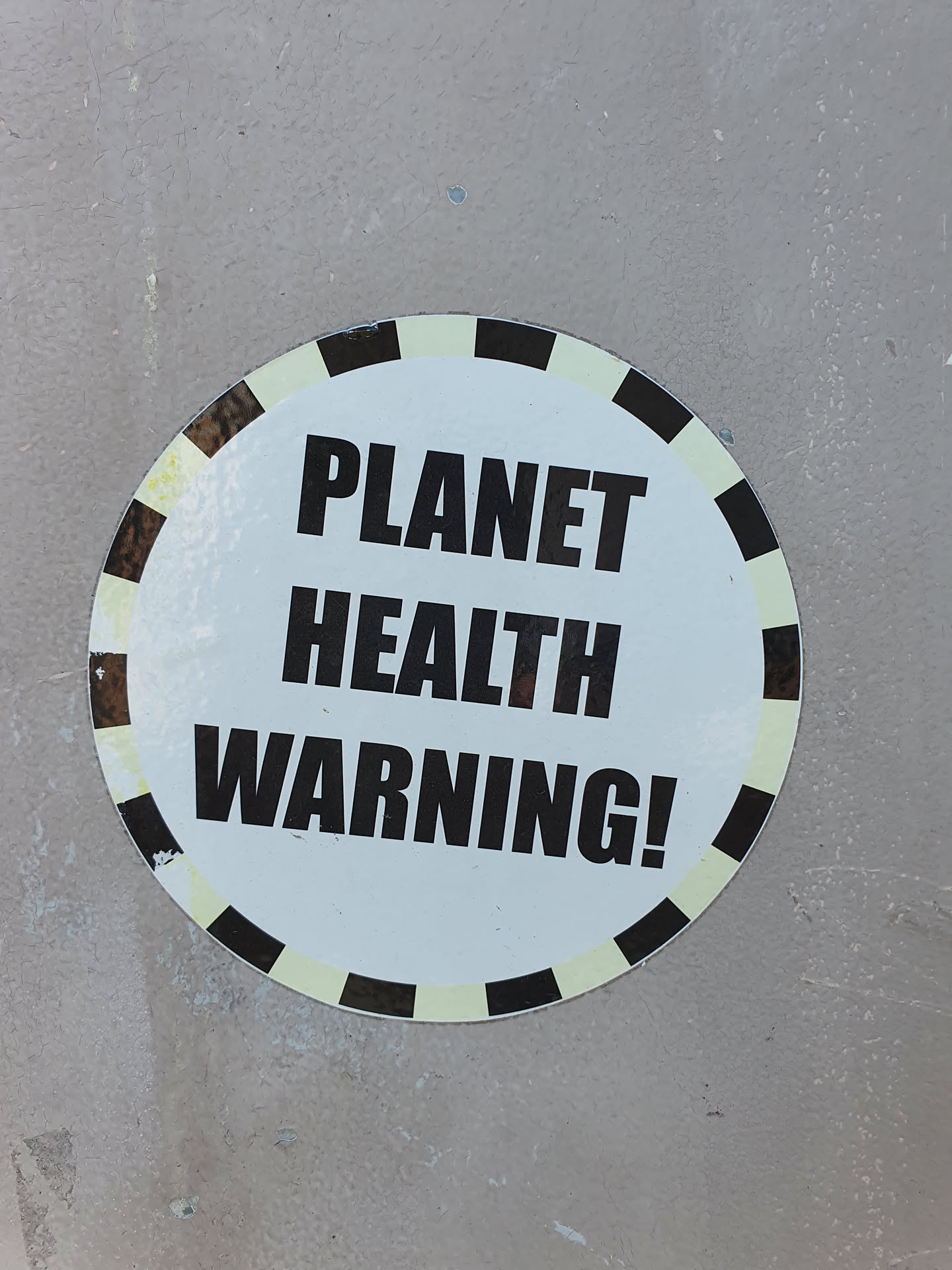 A round sticker with a black and white design.  The text 'PLANET HEALTH WARNING!' is prominently displayed in bold, black capital letters on a white background. The sticker has a decorative border of alternating black and off-white segments.