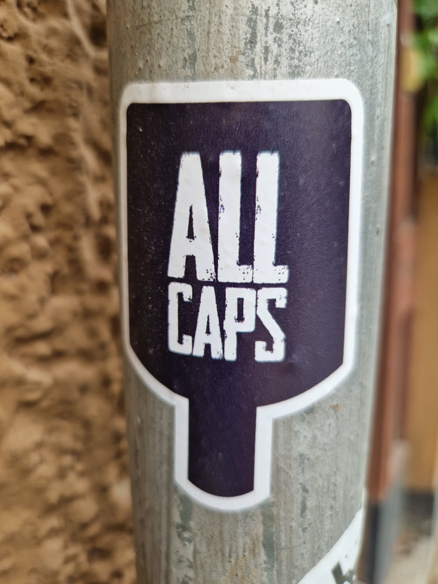 A sticker with the text "ALL CAPS" in white uppercase letters on a dark purple background. The sticker is shaped like a speech bubble with a bottom point. It is affixed to a weathered gray metal pole.
