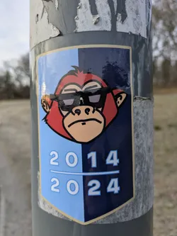 A sticker featuring a cartoon monkey wearing sunglasses. The sticker is split into two colors, light blue and dark blue, with the years 2014 and 2024 written below the monkey. The style is simple and somewhat reminiscent of 90s cartoons.