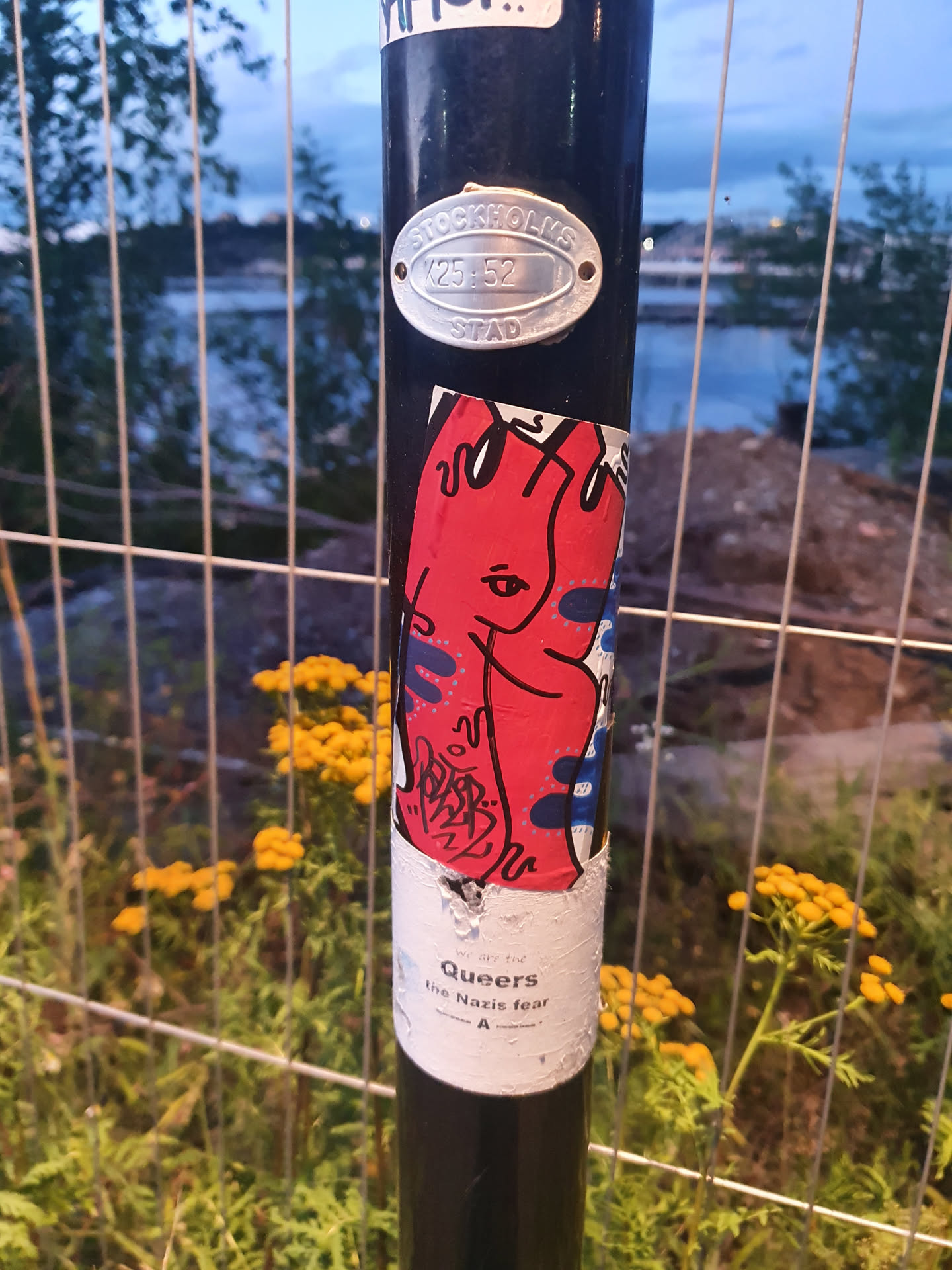 A sticker featuring an abstract figure in red and blue colors. The sticker is affixed to a dark-colored pole, surrounded by vegetation.  The text "Queers the Nazis fear" is visible below the main graphic.
