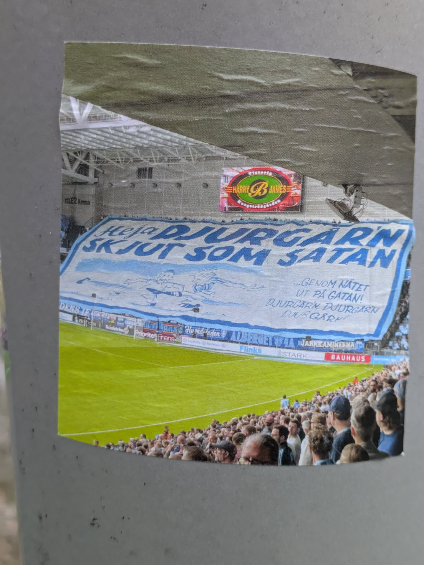 A sticker depicting a soccer stadium scene with a large banner reading "Heja Djurgården Skjut som Satan" (Go Djurgården! Shoot like Satan). The banner also includes additional text in Swedish. The sticker shows a lively match atmosphere with spectators in the stands.