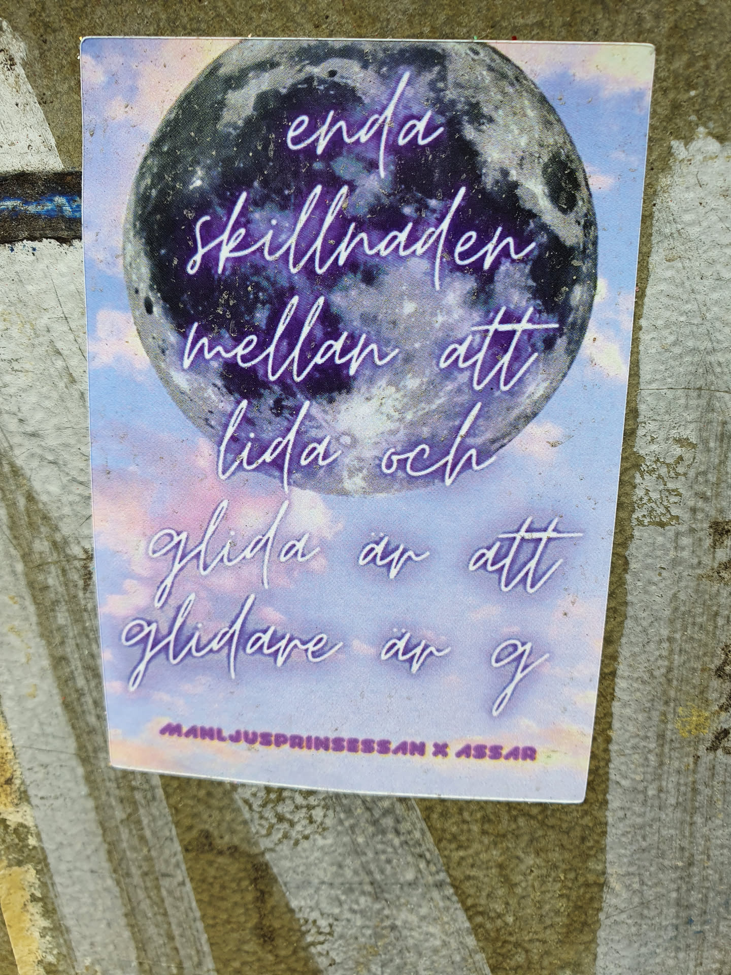 En rektangulär sticker med en bild av månen i bakgrunden. Texten på stickern är skriven på svenska och lyder: "enda skillnaden mellan att lida och glida är att glida är g". Längst ner på stickern står det: "MANLJUSP RINSSSAN X ASSAR".