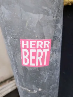 A pink sticker with the text "Herr Bert" on a gray metal surface.