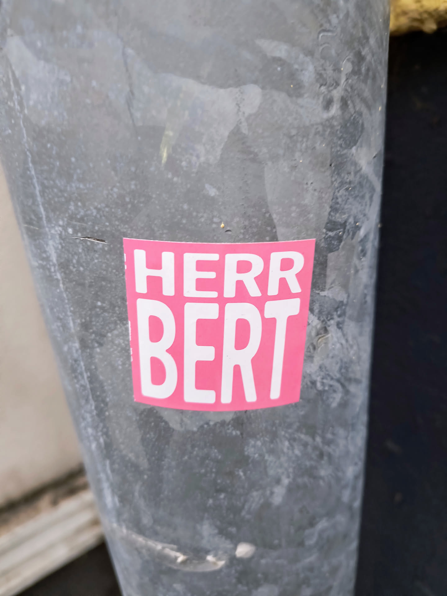 A pink sticker with the text "Herr Bert" on a gray metal surface.