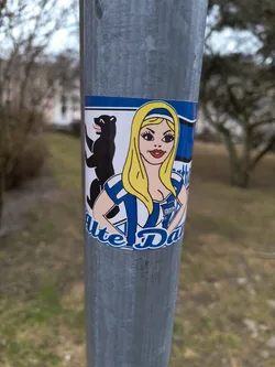 A sticker featuring a cartoon illustration of a blonde woman in a blue and white uniform, alongside a bear. The text 'Alte Dame' is visible at the bottom. The sticker is affixed to a metal pole outdoors.