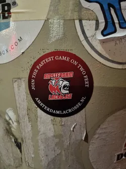 Round sticker featuring the Amsterdam Lacrosse logo. The logo is a red and white illustration of a lacrosse player's head, with the team name and website address printed underneath.  The sticker also features the phrase "Join the Fastest Game on Two Feet." 