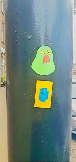 Two stickers are placed vertically on a metal lamppost. The top sticker is a cartoonish, light green creature with large yellow eyes and a triangular orange beak. It is set against a clear circular background. Below it is a smaller rectangular sticker with a yellow background. On the yellow background is a blue, pixelated shape that resembles a creature or object. The pole has a dark, glossy finish.