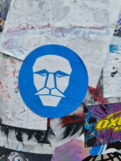 A round blue sticker with a white geometric face design. The face is simplistic with sharp lines and a mustache.