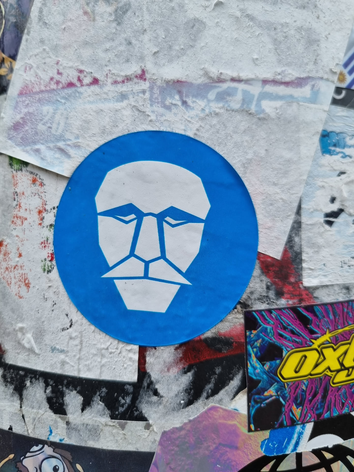 A round blue sticker with a white geometric face design. The face is simplistic with sharp lines and a mustache.