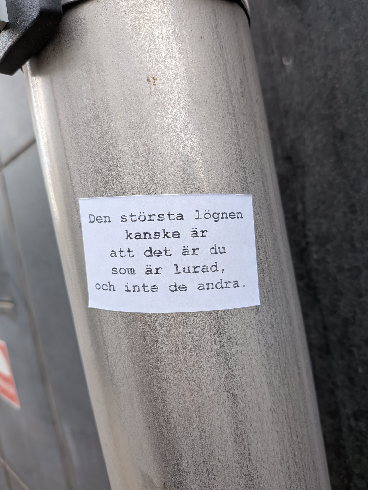 A rectangular white paper sticker with black text printed in a typewriter-style font. The sticker is adhered to a brushed metallic cylindrical surface, possibly a pole or a container. The edges of the paper appear slightly worn or curled, indicating it's a physical object. The text is centered on the sticker.
