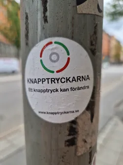 A round sticker with a logo and text. The logo is a circle with three colored segments. The text says "KNAPPTRYCKARNA" and "Ett knapptryck kan förändra" which translates from Swedish to "One button press can change".  There is also a website address at the bottom.