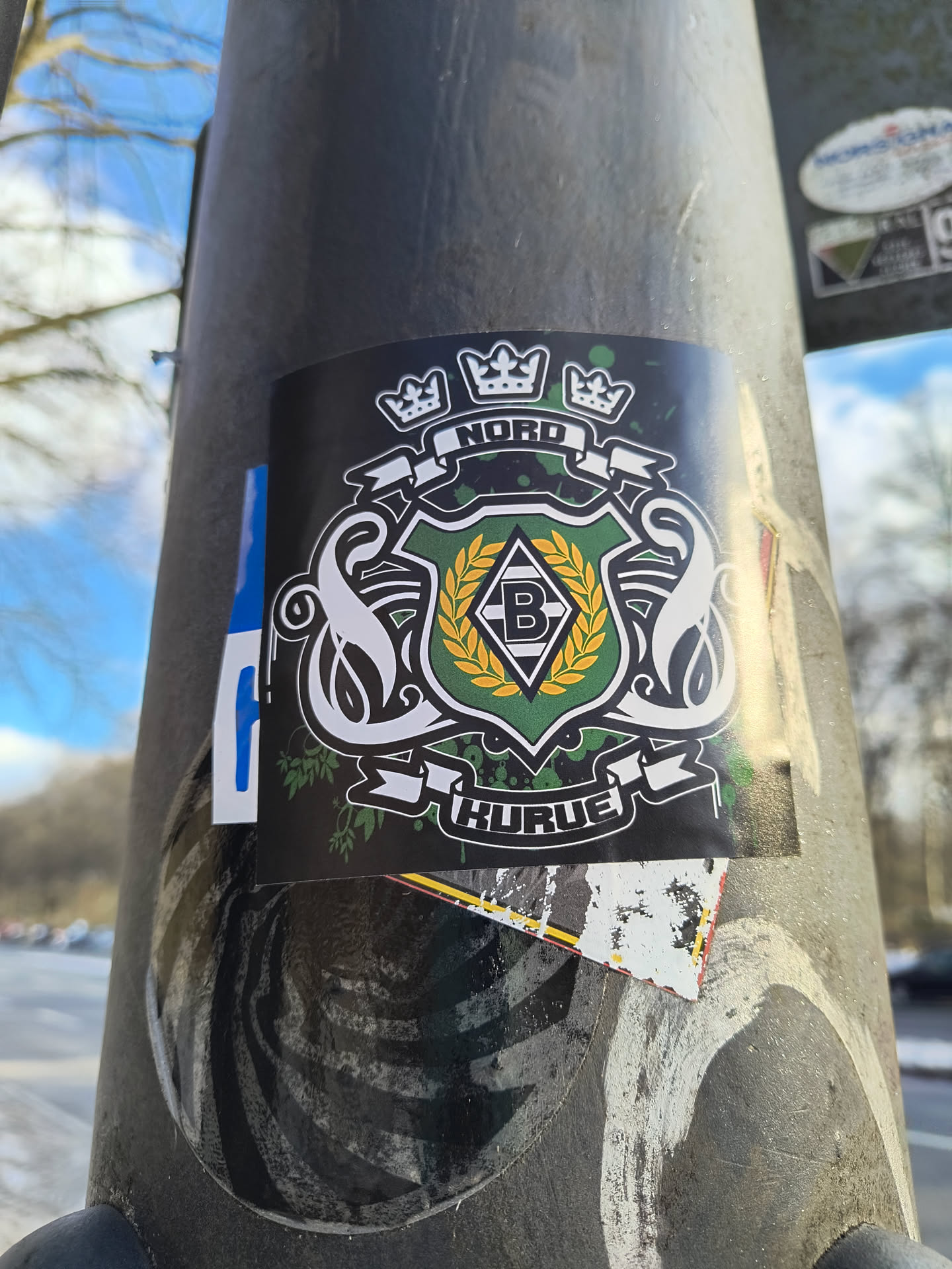 This is a black sticker on a metallic post featuring the Borussia Mönchengladbach logo. The logo has a black background with a green and white shield in the center containing a black 'B' on a white diamond. The shield is surrounded by a gold laurel wreath and flanked by two white swans. Above the shield are three crowns, and banners above and below read 'NORD' and 'KURVE' respectively.