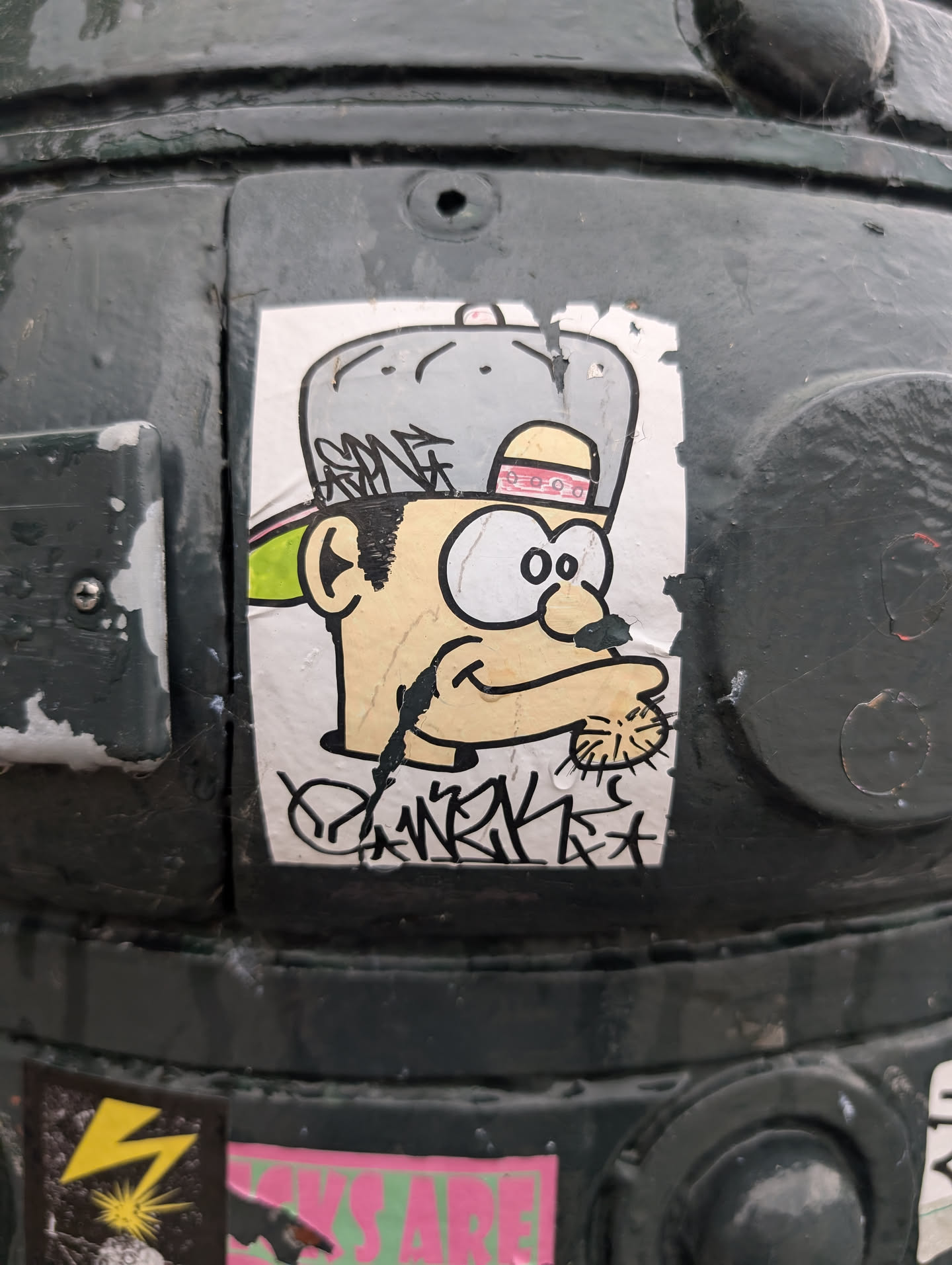 The sticker displays a stylized cartoon character's head against a white background. The character has large, expressive eyes with black pupils and white scleras, a small, pointed nose, and a wide, curved mouth. It is wearing a large, grey baseball cap with a pink and red band around the brim, and a green accent is visible behind its ear. The character's skin tone is light yellow, and its hair is depicted with simple black lines. Below the character's chin, there is a black drip line extending downwards, and across the bottom of the sticker is black graffiti-style lettering. The sticker appears to be slightly worn and adhered to a dark, textured surface.