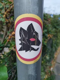 A worn-out AS Roma Lupetto (wolf cub) sticker affixed to a metallic pole.  The sticker shows the iconic black wolf's head on a white background, encircled by red and gold rings.  The sticker appears weathered, showing scratches and some discoloration.