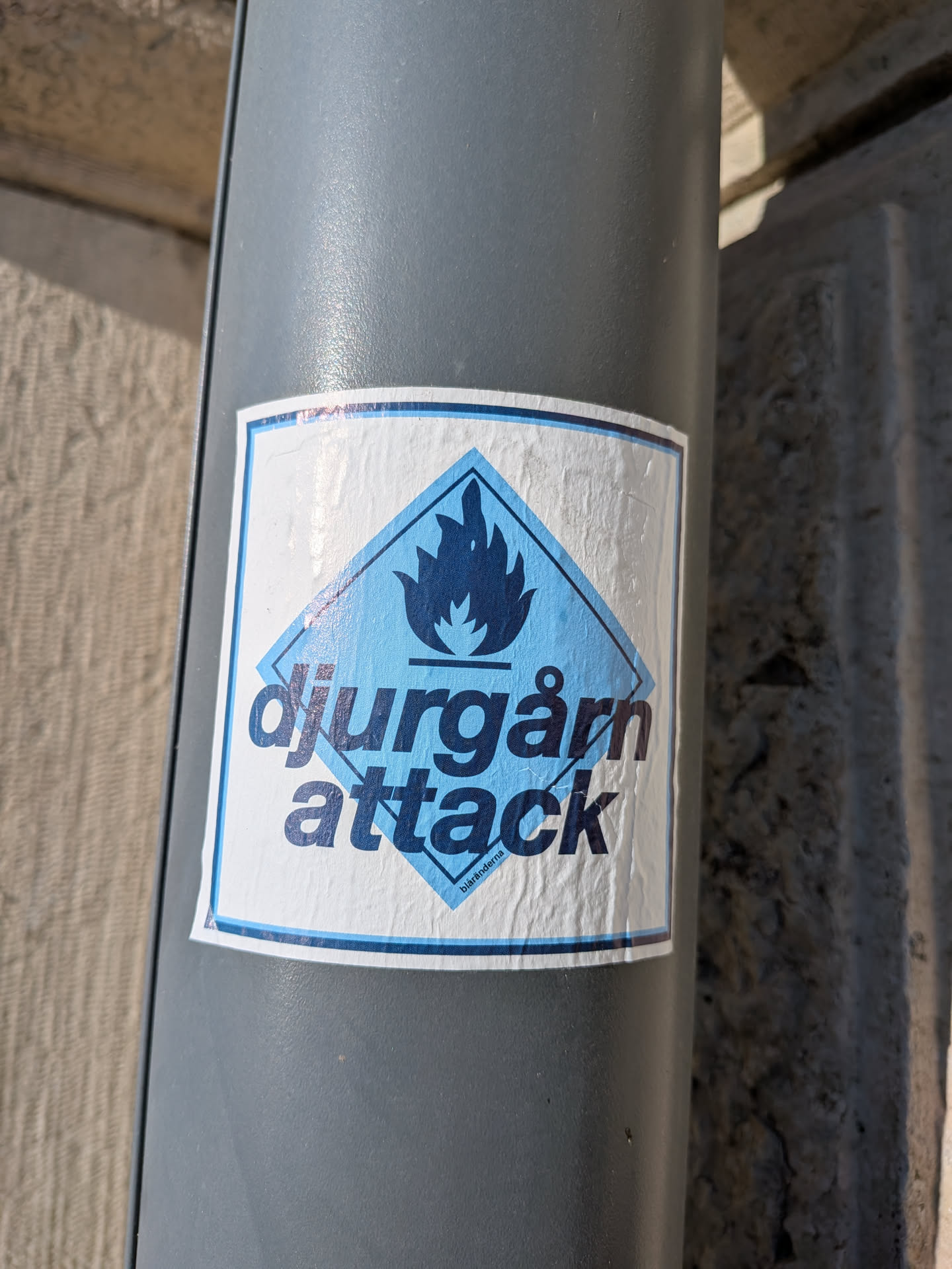 The sticker is rectangular with a white background and a blue border. At the center, there is a blue diamond shape containing a stylized blue flame symbol. Below the flame, the text 'djurgårn attack' is written in large, dark blue, sans-serif capital letters. At the very bottom of the diamond, in smaller text, are the words 'biaränderna'. The sticker appears slightly creased and aged, suggesting it is a real sticker applied to a surface.