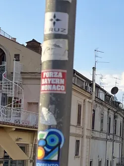 Street sticker The sticker is rectangular with a bright red background and bold white capitalized text. The text is arranged in three lines: 'FORZA' on the top, 'BAYERN' in the middle, and 'MONACO' at the bottom. The sticker appears slightly worn around the edges, indicating it's a physical adhesive item on a grey pole.