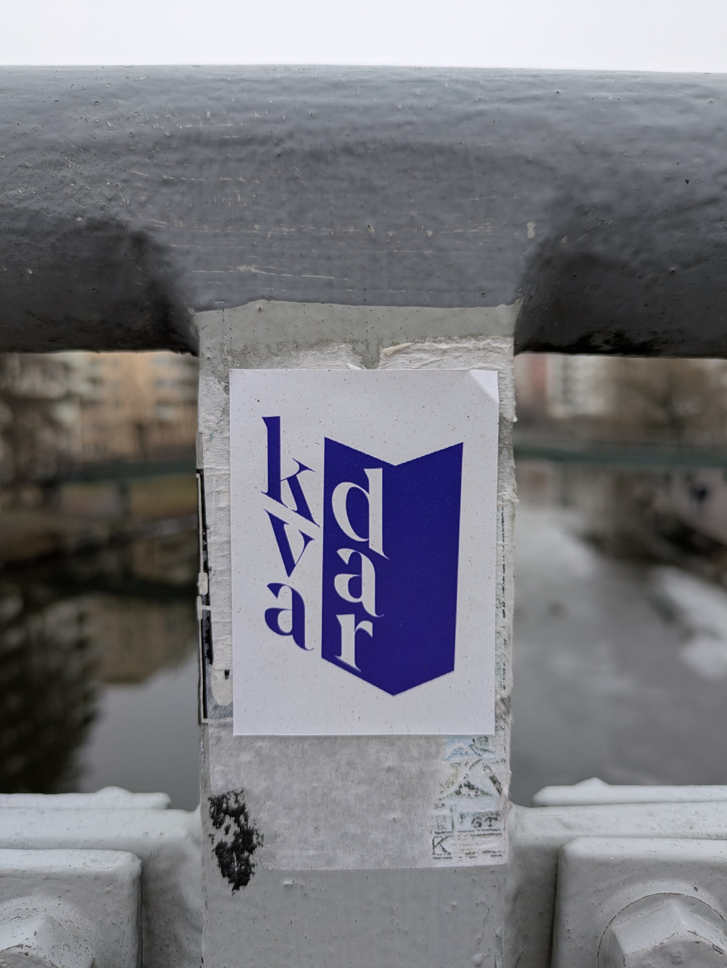 A white sticker with the word "kdaar" in blue, attached to the side of a bridge. The text is oriented vertically. The bridge is gray and overlooks a body of water. The sticker is simple and clean in design.