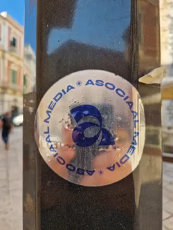 A silver circular sticker with the text "SOCIAL MEDIA * ASSOCIAL MEDIA" repeating around the edge.  A stylized blue logo is in the center. The sticker is affixed to a dark-colored metal pole.