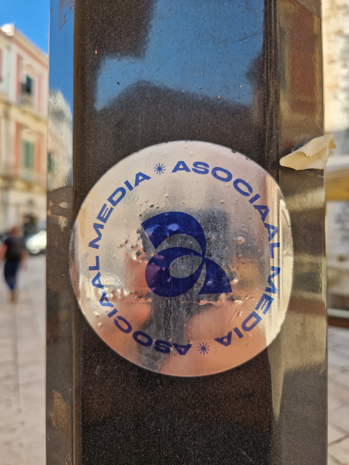 A silver circular sticker with the text "SOCIAL MEDIA * ASSOCIAL MEDIA" repeating around the edge.  A stylized blue logo is in the center. The sticker is affixed to a dark-colored metal pole.