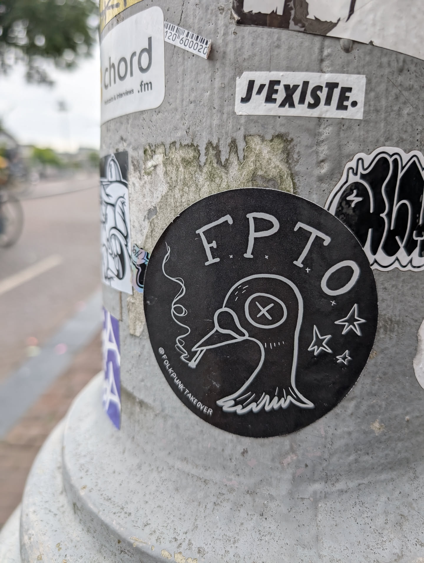 The sticker is circular and predominantly black with white line art. It depicts a bird with its head tilted, smoking a cigarette, and has 'X' marks for eyes. Several small stars are scattered around the bird. The text 'FPTO' is prominently displayed in a stylized font above the bird, and '@FOLKPUNKTAKEOVER' is written in smaller text along the bottom curve of the sticker. The overall aesthetic is edgy and hand-drawn.