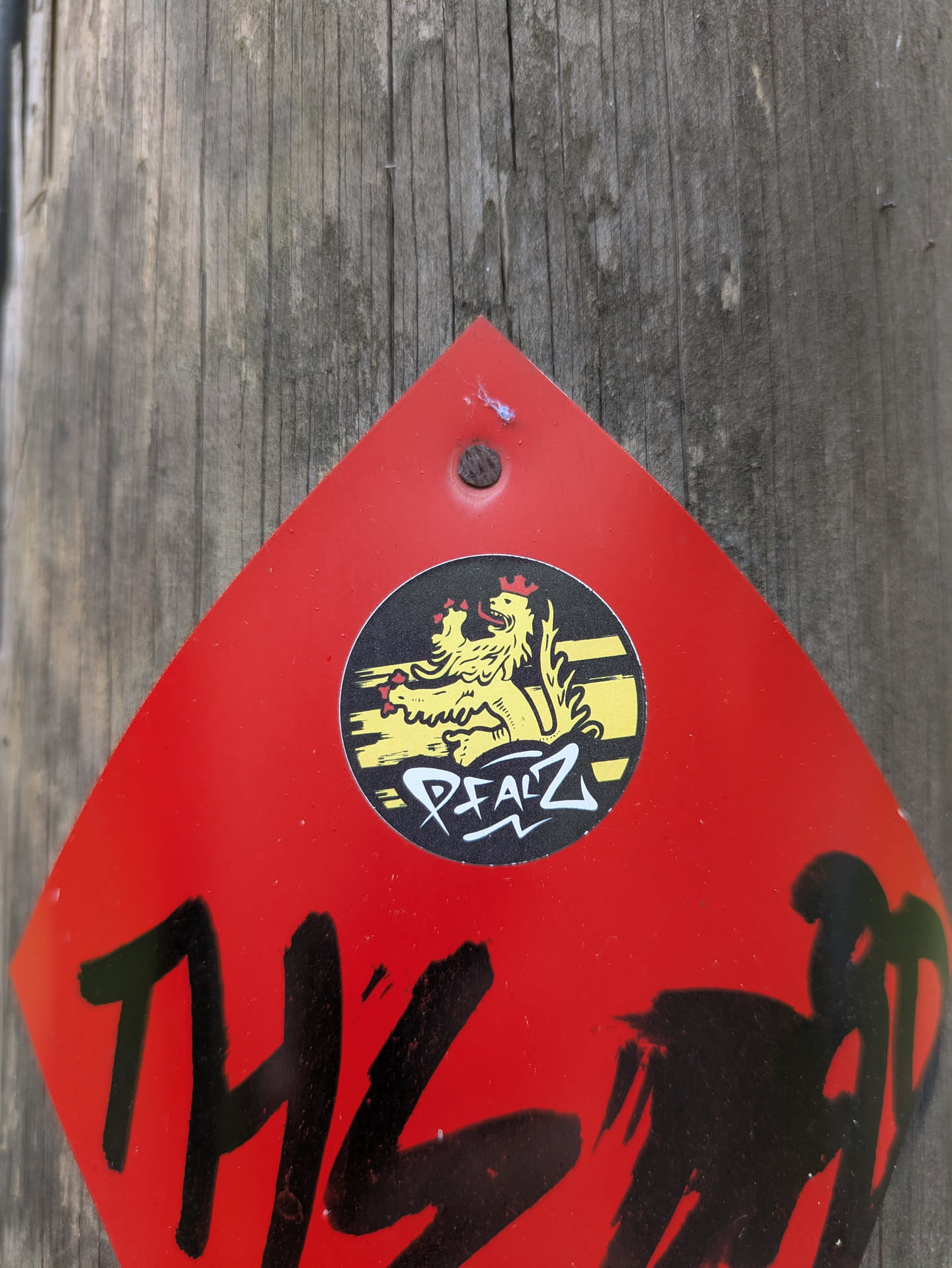 The image showcases a vibrant red, roughly diamond-shaped sign or large sticker, securely affixed to a weathered wooden pole with a single nail at its top. Centrally positioned on this red background is a circular black and yellow sticker. This inner sticker prominently displays a stylized yellow lion, adorned with a red crown and a red tongue, depicted in a rampant posture. Below the lion, the word "PFALZ" is written in a distinctive white, graffiti-style font with a black outline, against the black circular background. Further down on the main red sign, large, black, brushstroke-like letters are partially visible, appearing to spell "THIS" or a similar word, although the phrase is cut off by the bottom edge of the image.