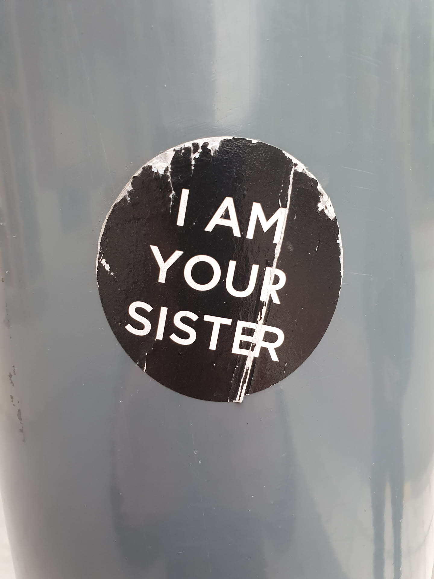 A round black and white sticker with the text "I AM YOUR SISTER" on it. The sticker is attached to a gray surface and is slightly damaged.