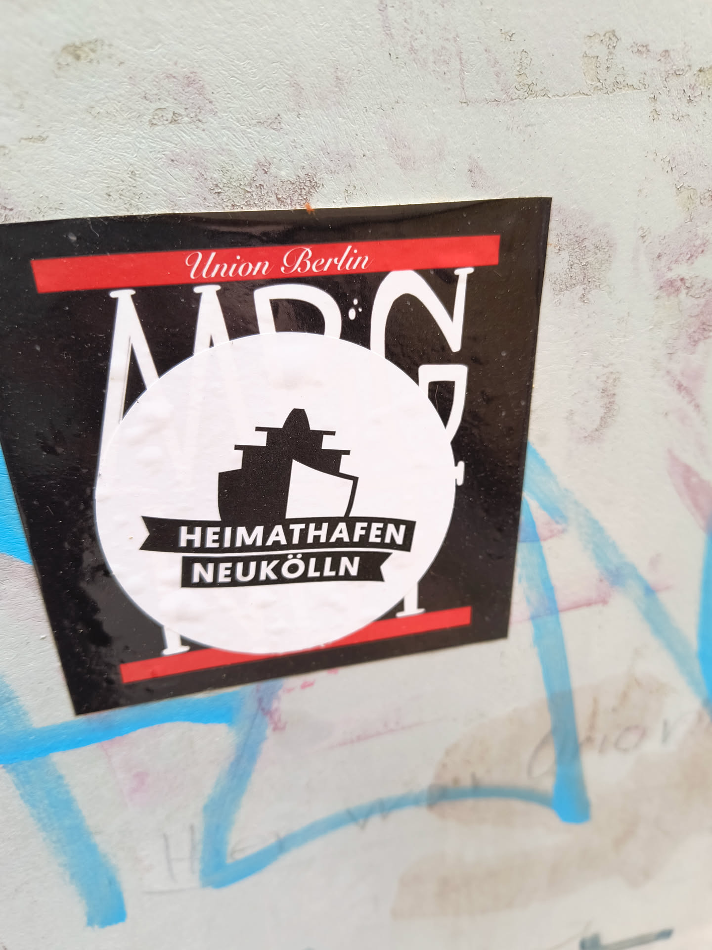 A black and white sticker with a red band at the top and bottom. The sticker features the text "Union Berlin" at the top, and a logo with a ship and the text "HEIMATHAFEN NEUKÖLLN" in the center. The background is black, and the text and logo are white.